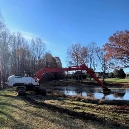 Excavator working in pond, water splashing, trees with fall foliage in background.