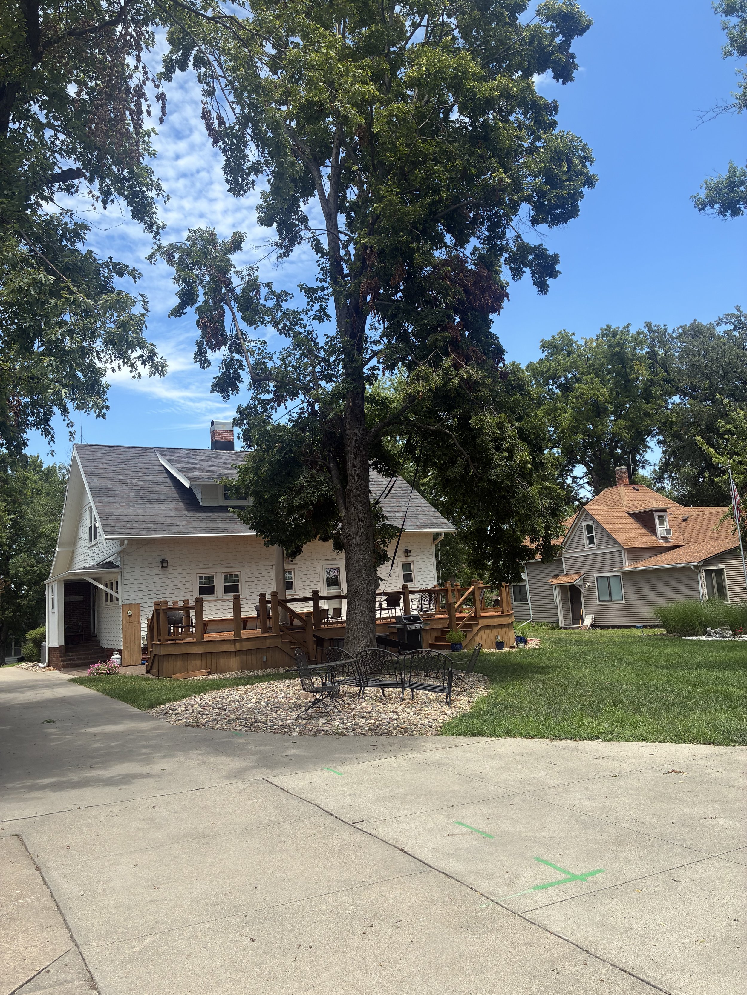 A suburban backyard with a large tree, a white house with a deck, and some outdoor furniture including a black metal bench and chairs. The yard has a grassy area and a section with decorative rocks, with some scaffolding and construction markings on the concrete patio.