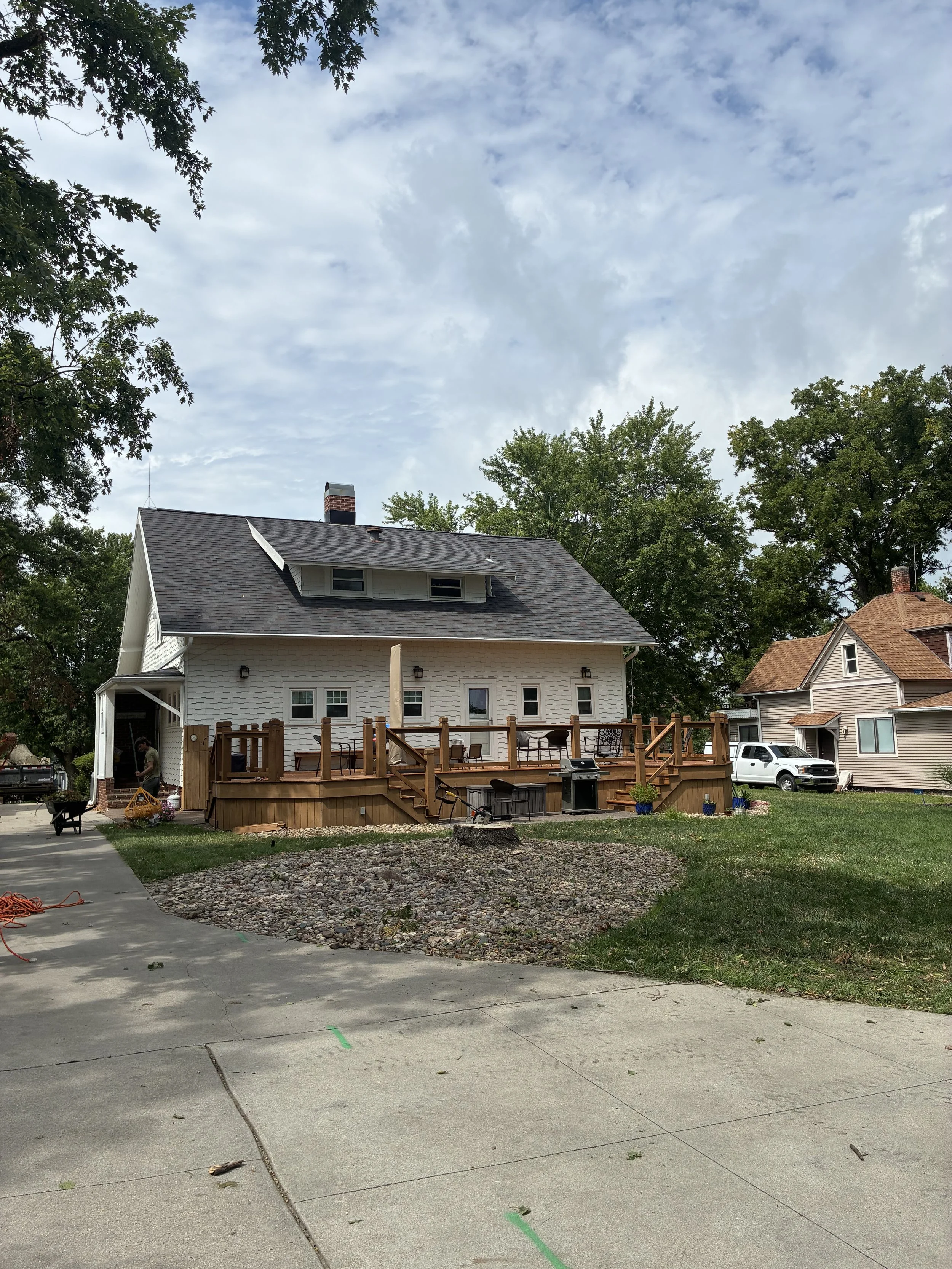 A white house with a gray roof and a chimney, under a partly cloudy sky, with a wooden deck in the backyard, surrounded by trees and neighboring houses, and a driveway with some green markings.