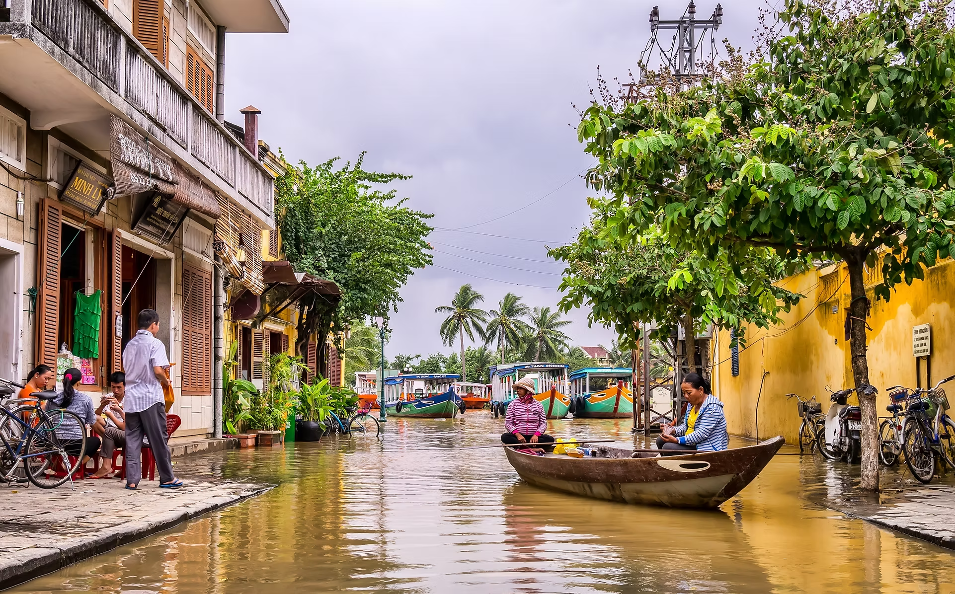 Rue de ville inondée avec des personnes en bateau, arbres verts, bâtiments avec fenêtres à volets, et des bateaux de touriste. Des individus sont assis ou se tiennent près des bâtiments, pendant que d'autres surfent ou naviguent à travers l'eau.