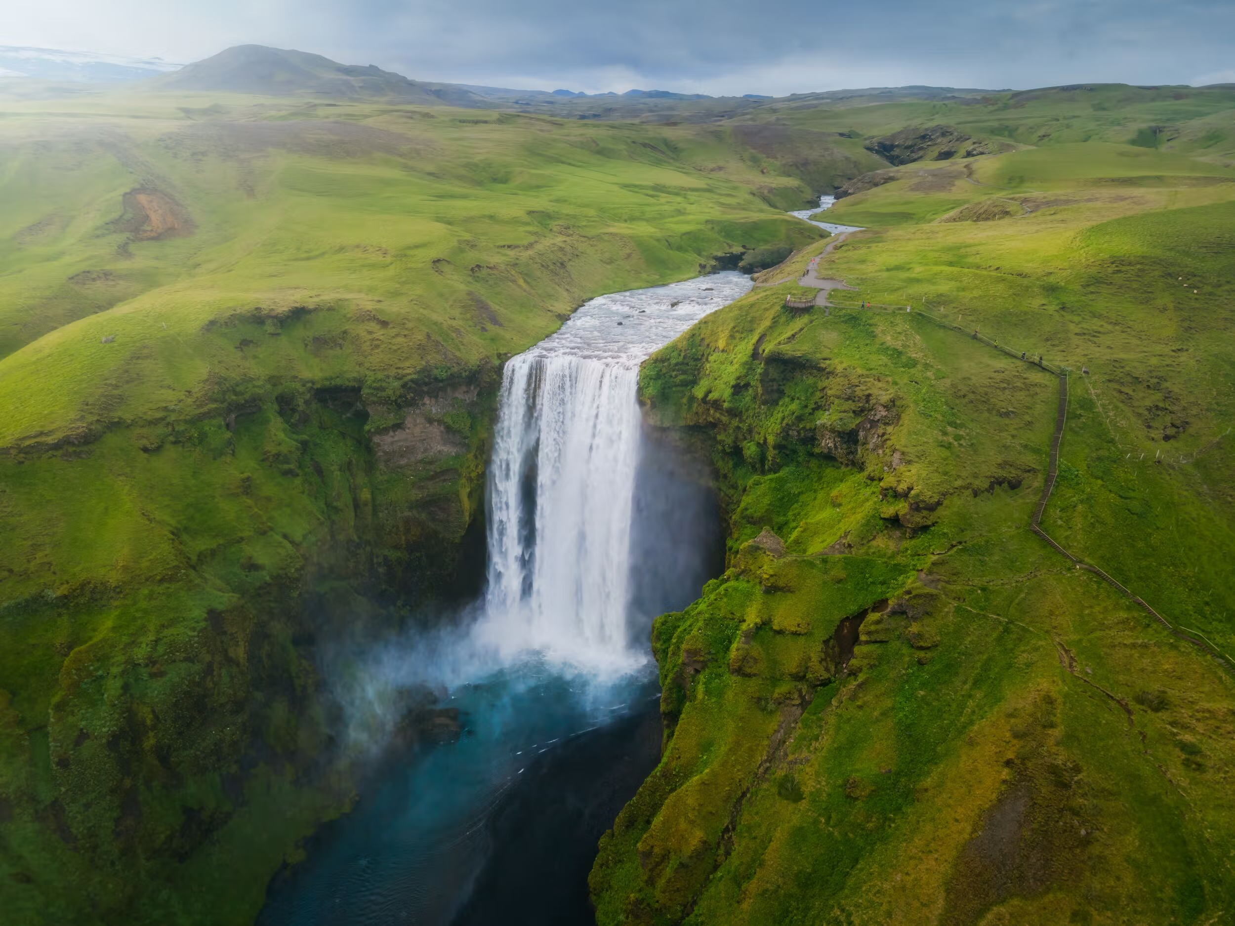 Cascade d'une rivière entourée de collines verdoyantes sous un ciel nuageux.