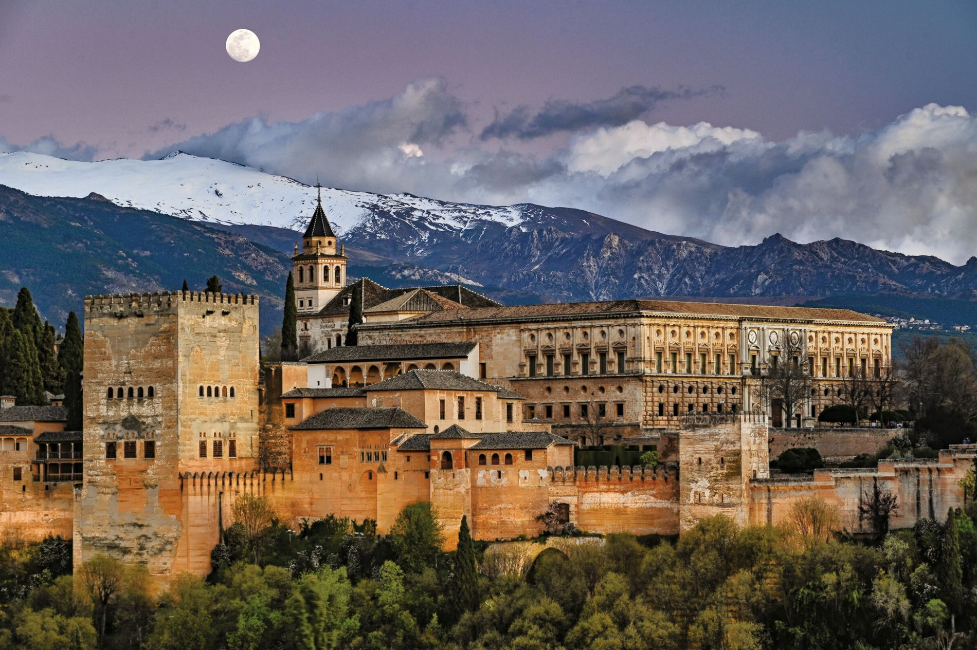 Une vue panoramique du Alhambra, un palais et forteresse historiques à Grenade, Espagne, avec la chaîne de montagnes de la Sierra Nevada en arrière-plan, sous un ciel semi-ombragé avec la pleine lune visible.