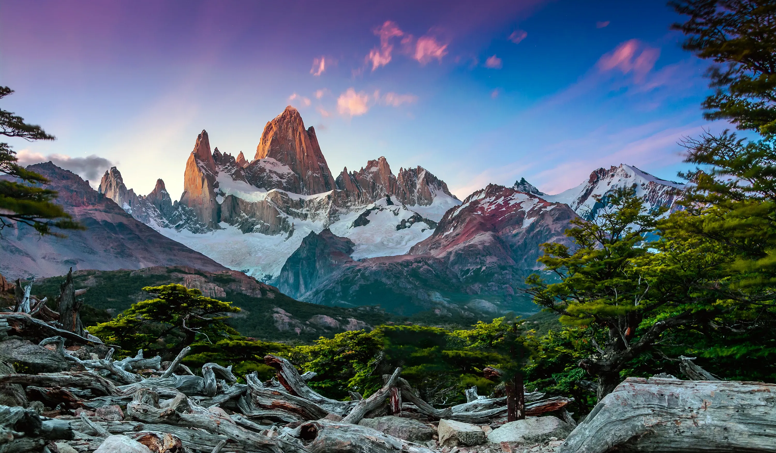 Montagnes enneigées et rocheuses sous un ciel bleu avec des nuages roses, entourées de forêt verdoyante.