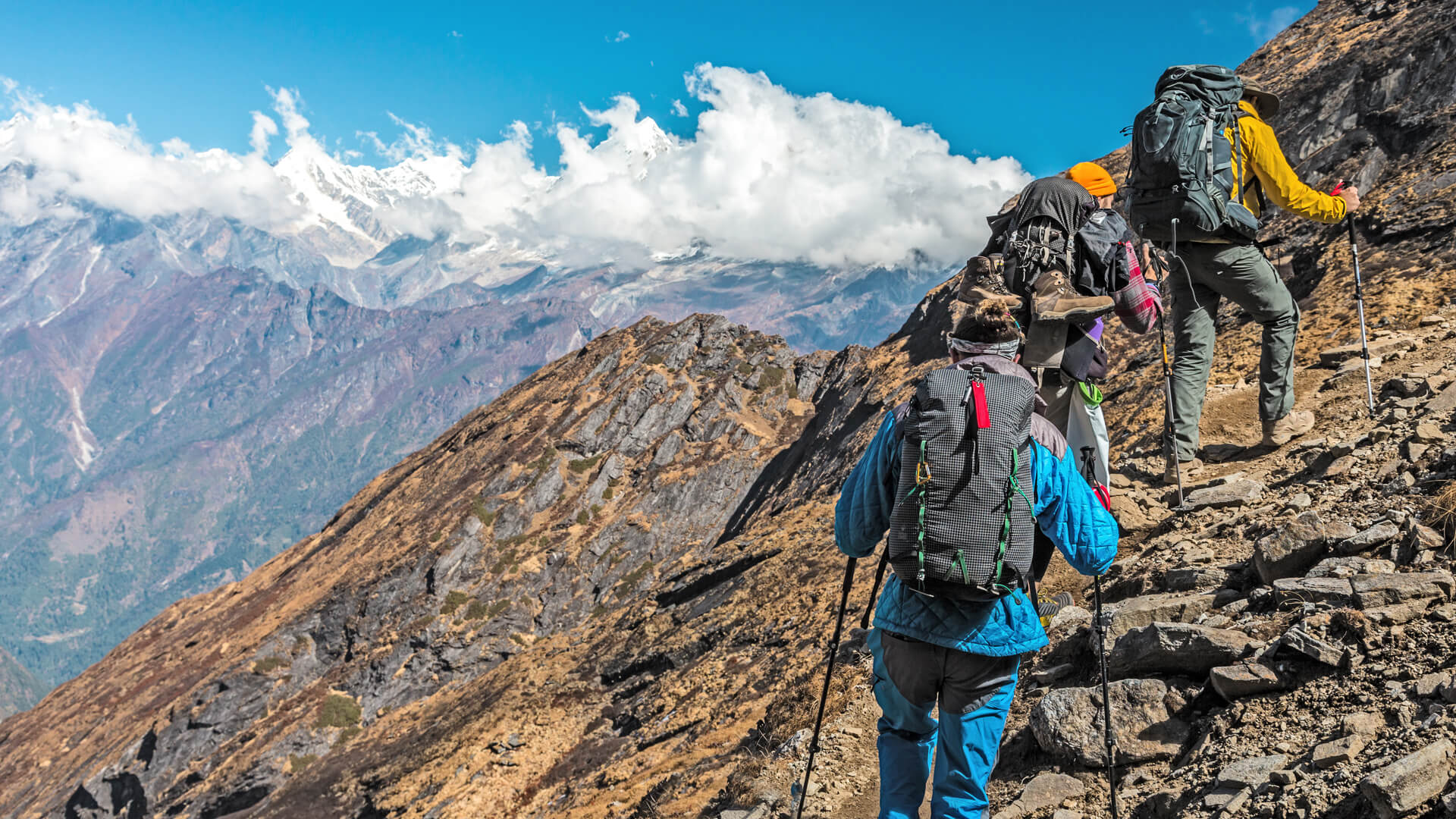 Groupe de randonneurs escaladant une montagne rocheuse avec des sommets enneigés en arrière-plan, sous un ciel bleu.