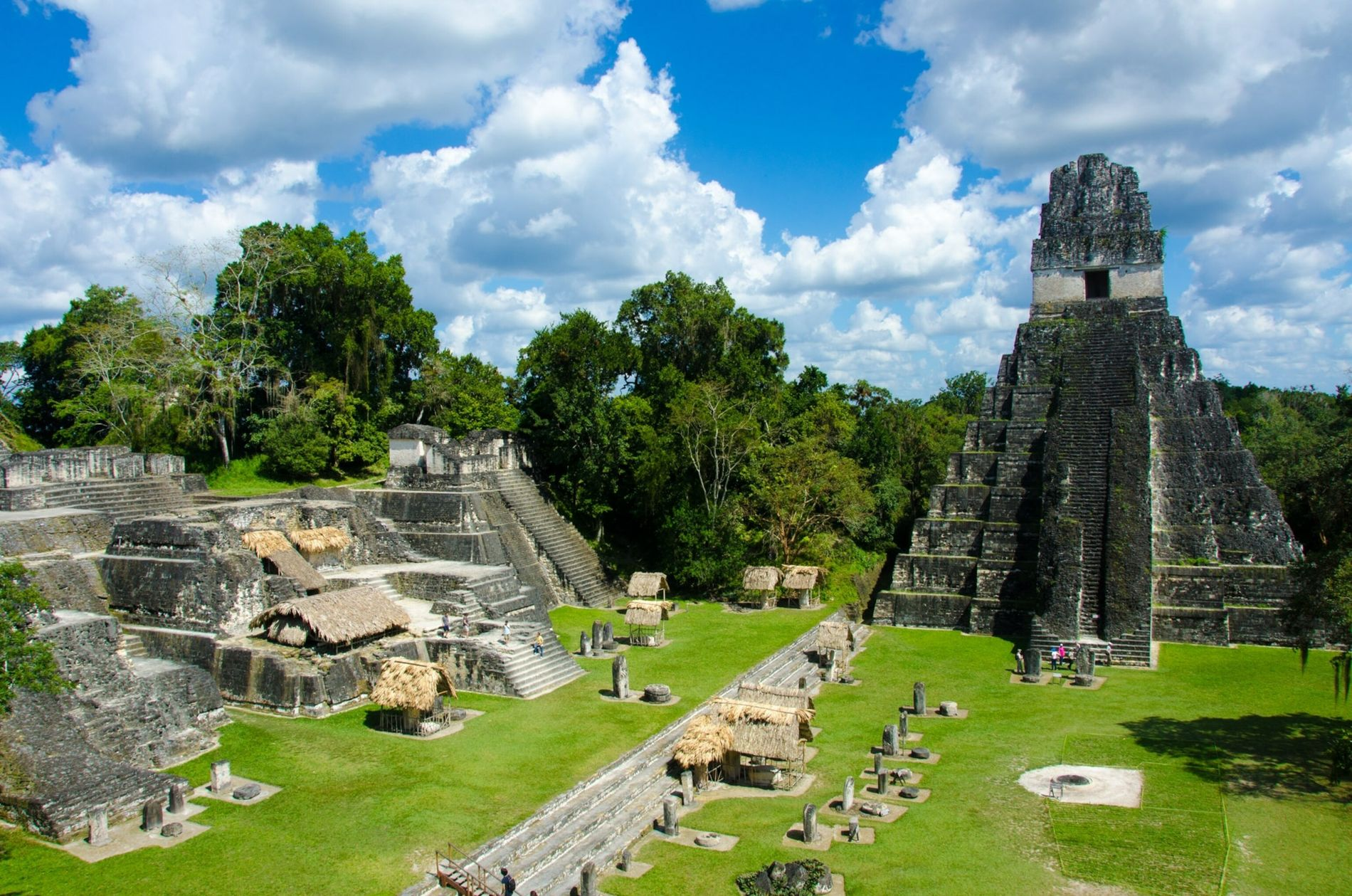 Ruines d'une ancienne cité maya avec une grande pyramide en pierre, entourée de structures plus petites et de huttes en chaume, dans une jungle verdoyante sous un ciel en partie nuageux.