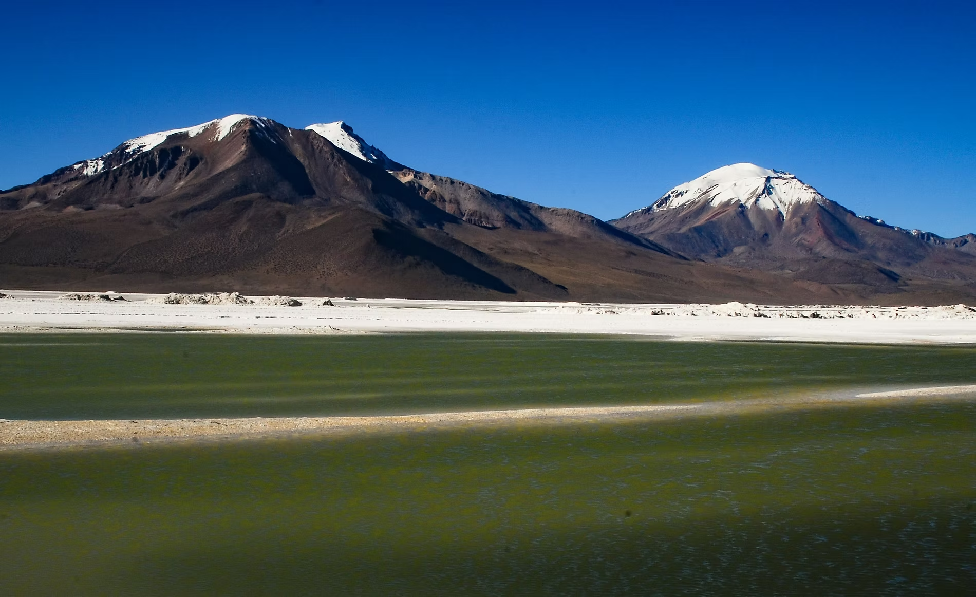 Deux montagnes avec des sommets enneigés, un paysage aride au premier plan, et un ciel bleu clair.