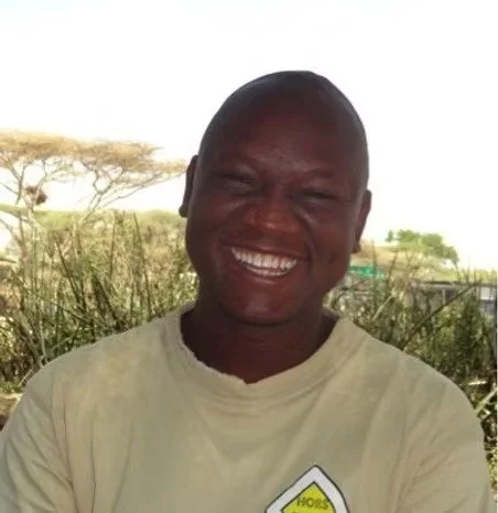 Une personne souriante, de peau foncée, portant un t-shirt beige, en plein air avec des arbres en arrière-plan.