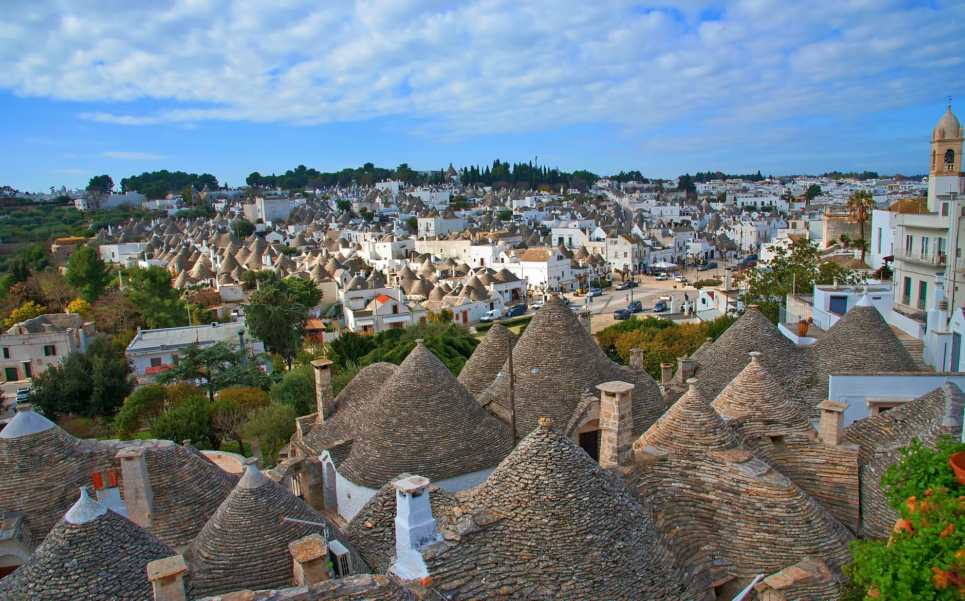 Vue aérienne des toits en forme de cône traditionnels dans une ville blanche ensoleillée, avec un ciel nuageux, des bâtiments modernes et des espaces verts.