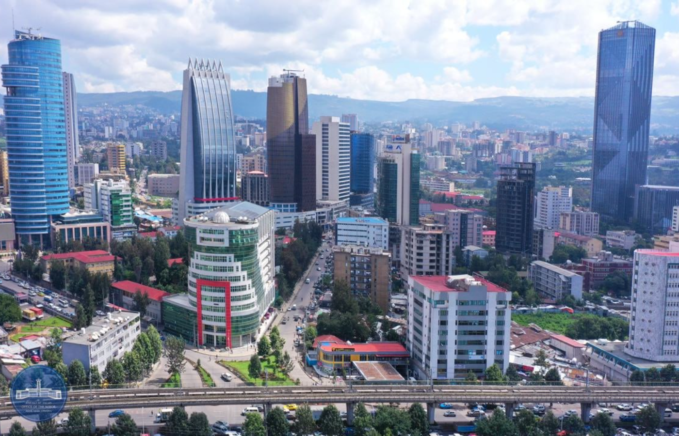Vue aérienne du centre-ville avec de nombreux gratte-ciel modernes, une autoroute en premier plan, des bâtiments résidentiels et commerciaux, le relief montagneux au loin, un ciel partiellement nuageux.