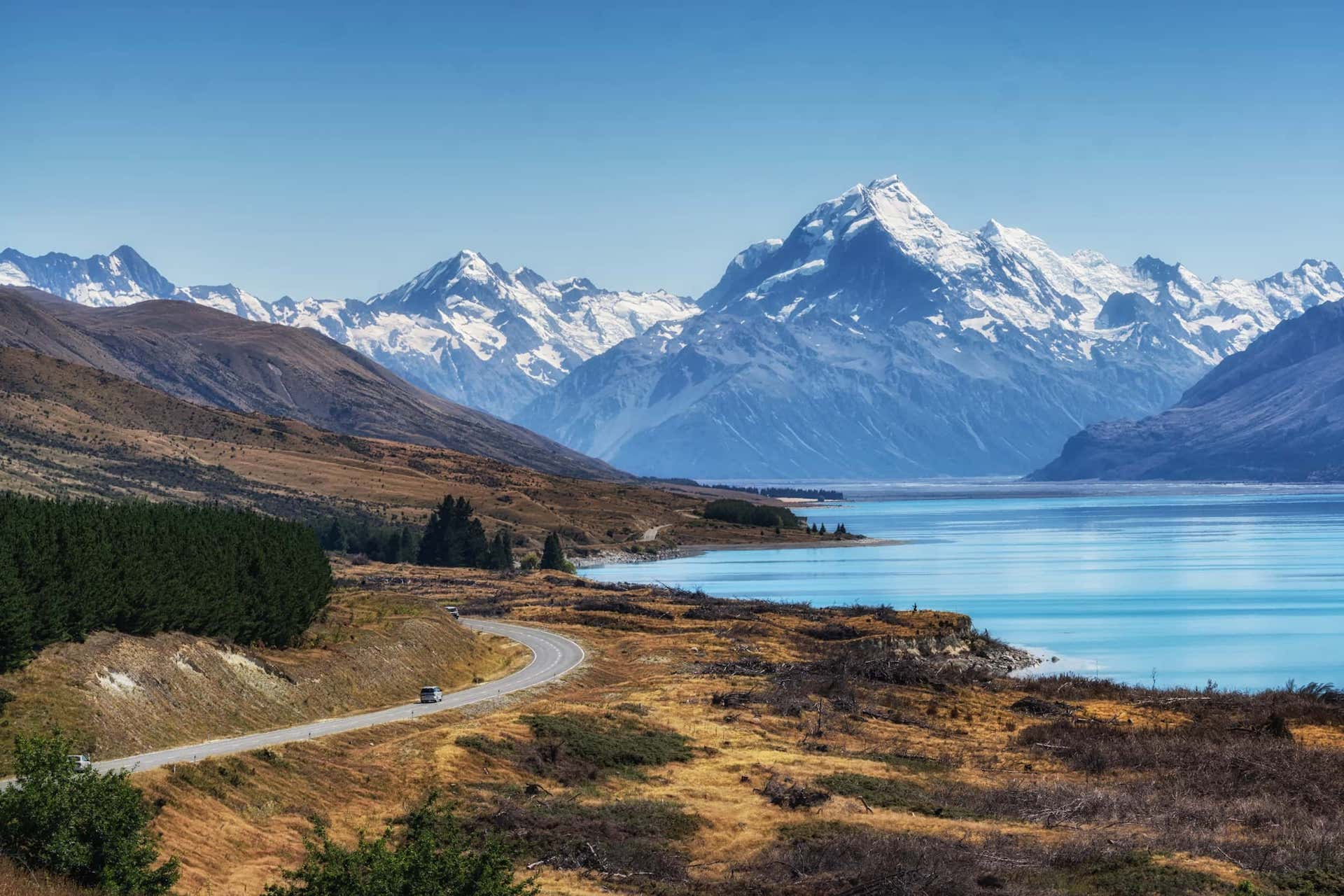 Paysage montagneux avec des montagnes enneigées, un lac bleu et une route serpentant à travers des terrains vallonnés avec de la végétation.