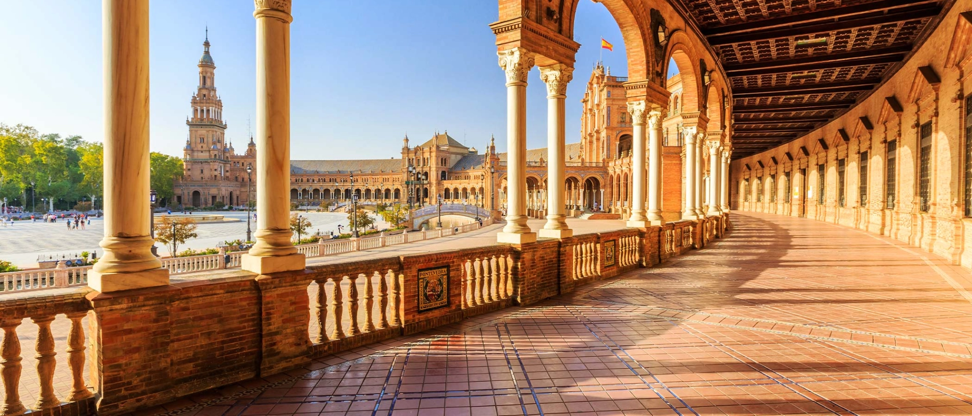 Vue d'une architecture historique avec des colonnes blanches et un balustrade en pierre, un grand bâtiment en arrière-plan avec une tour haute et un drapeau espaol, sous un ciel bleu.