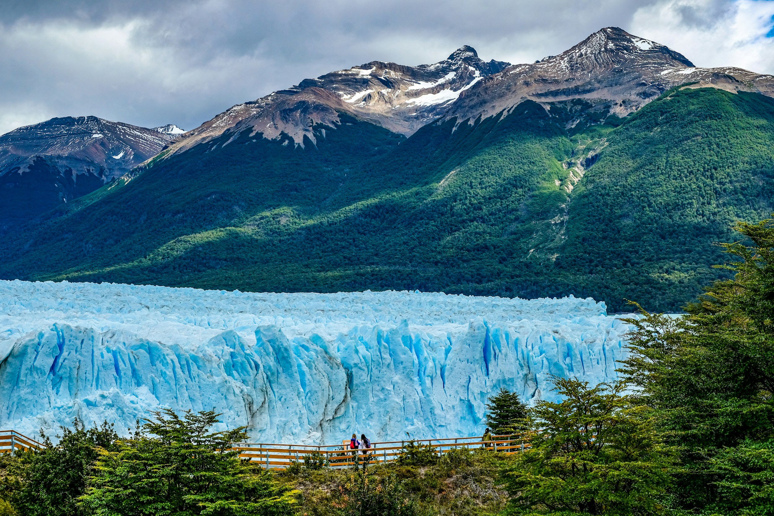 Paysage de montagnes avec glacier bleu en premier plan, forêt verte et sommets enneigés sous un ciel nuageux.