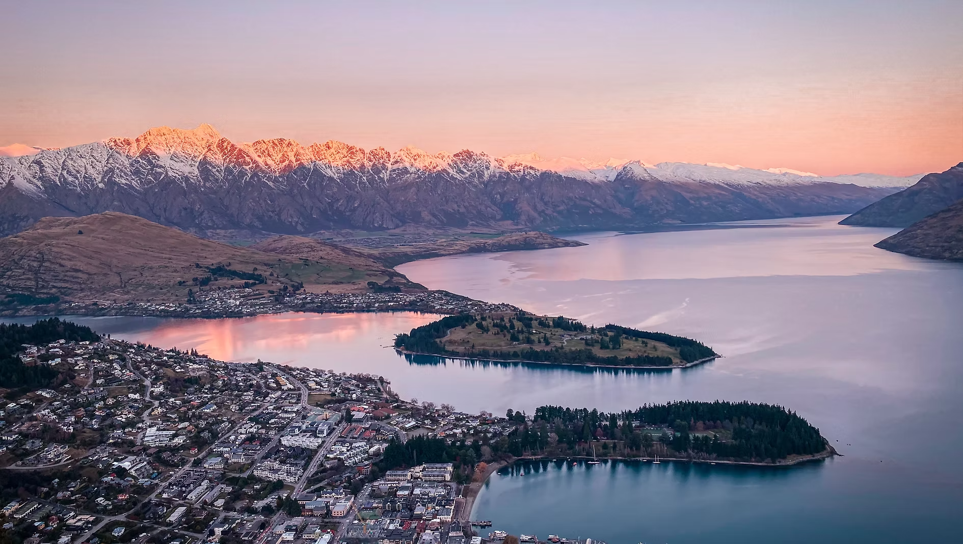Vue aérienne d'une ville au bord d'un lac entouré de montagnes enneigées au coucher du soleil, avec des reflets roses et oranges dans l'eau.