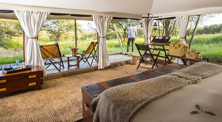 Intérieur d'une tente de luxe avec vue sur la nature, meubles en bois, un lit avec une couverture, et une personne regardant le paysage extérieur.