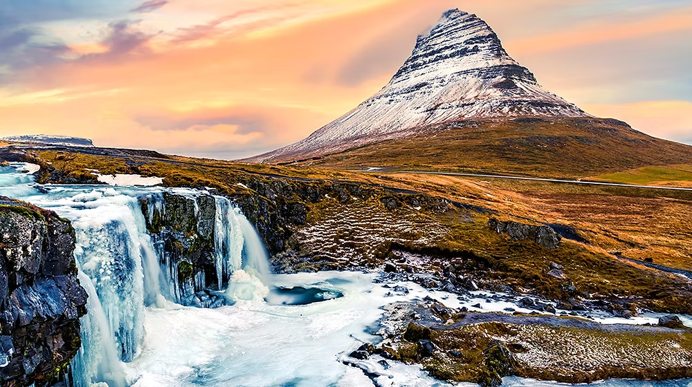 Paysage avec une cascade, un volcan enneigé en arrière-plan, ciel coloré au coucher du soleil, et terrain rocheux avec de la végétation