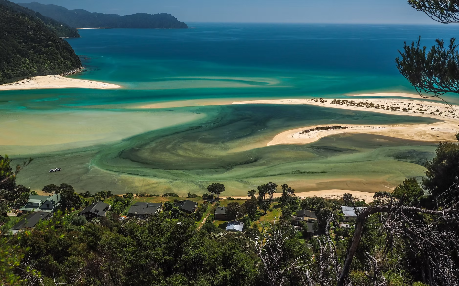 Paysage côtier avec une plage de sable, une mer turquoise, une forêt dense et un village résidentiel en contrebas.