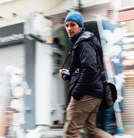 Jeune homme avec une barbe, portant un bonnet bleu, une veste noire, un sac à dos noir, marchant dans une rue urbaine.