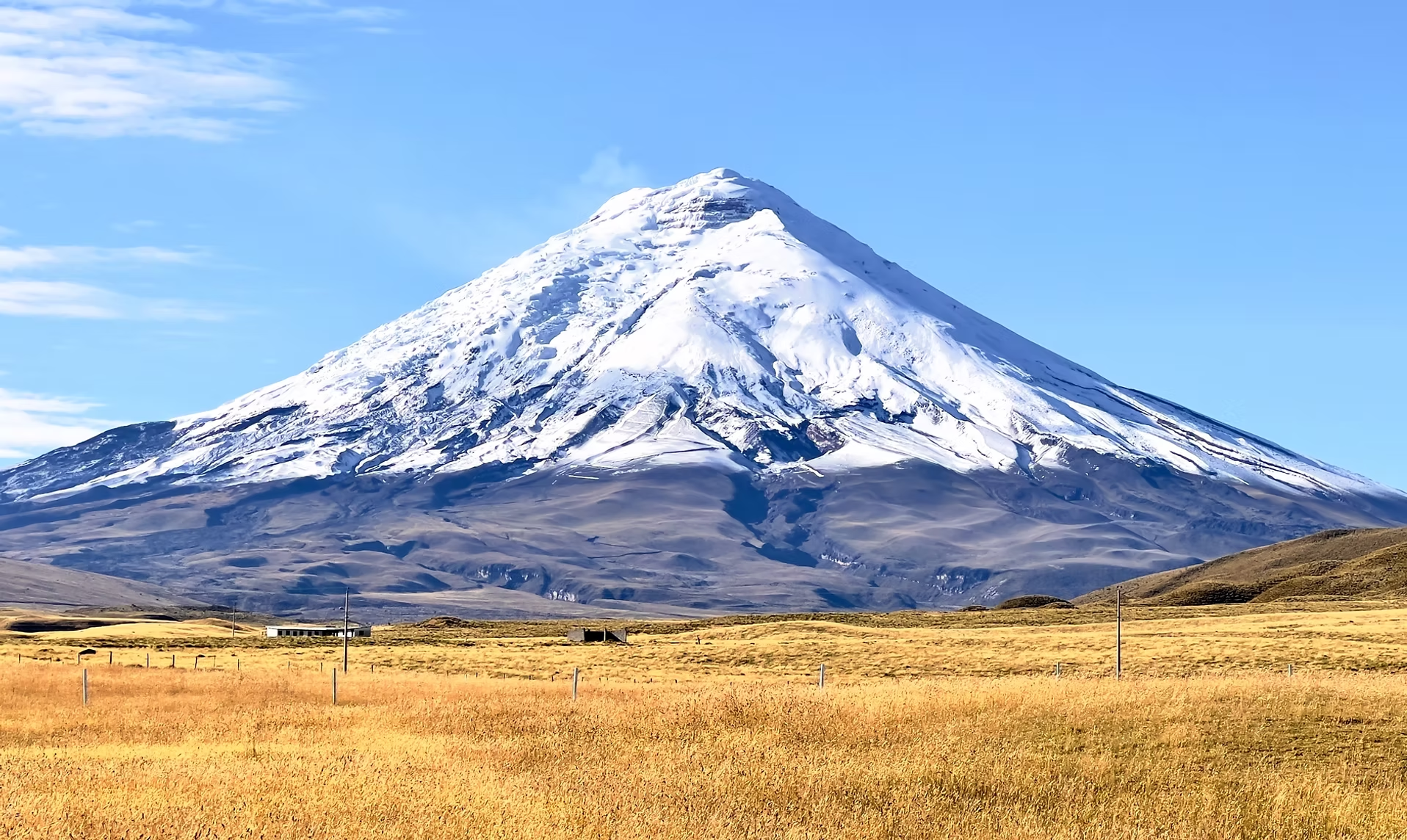 Montagne enneigée avec sommet volcanique, paysage pastoral avec prairies et quelques bâtiments au premier plan.