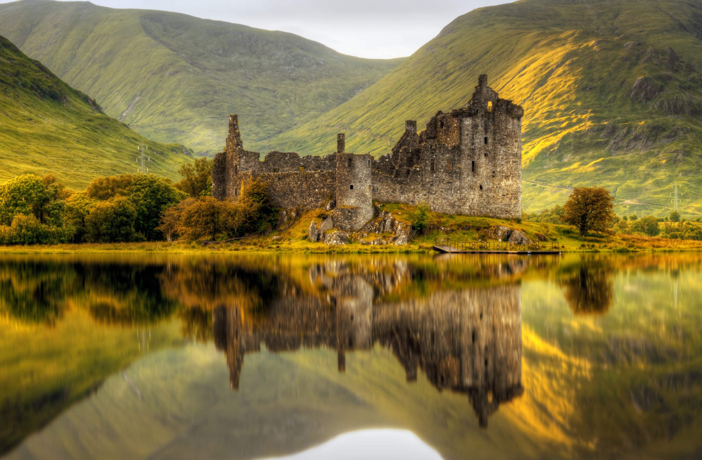 Château en ruines au bord d'un lac entouré de collines verdoyantes et montagnes, reflet dans l'eau.