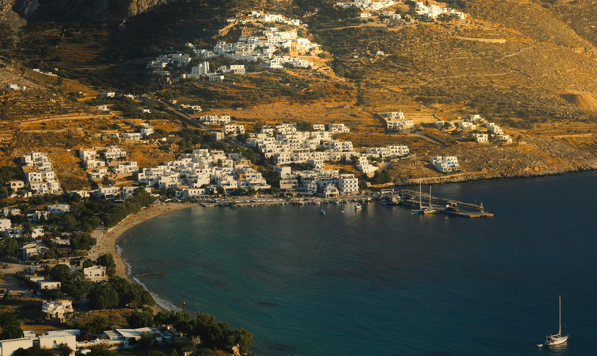 Vue aérienne d'un village côtier avec des maisons blanches sur une pente rocheuse et un port avec des bateaux, au bord d'une mer calme, au coucher du soleil.