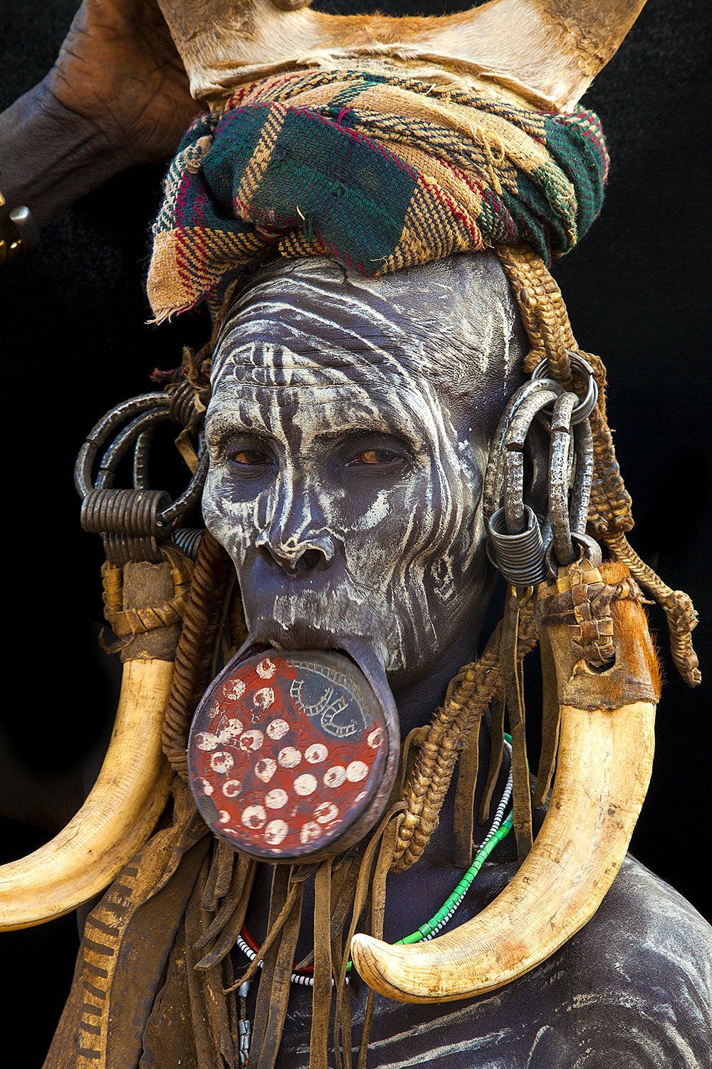 Une personne avec une peau peinte en motifs noirs et blancs, portant de grands anneaux en métal dans les oreilles, un collier de perles et un couvre-chef en tissu tartan, qui porte un masque facial circulaire avec des motifs blancs. Elle a également des grandes cornes en os ou en ivoire autour du cou.