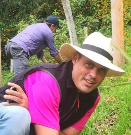 Deux hommes travaillant dans un jardin ou un champ, dont un portant un large chapeau blanc avec une bande noire, et l'autre portant une casquette, entourés de végétation verdoyante.
