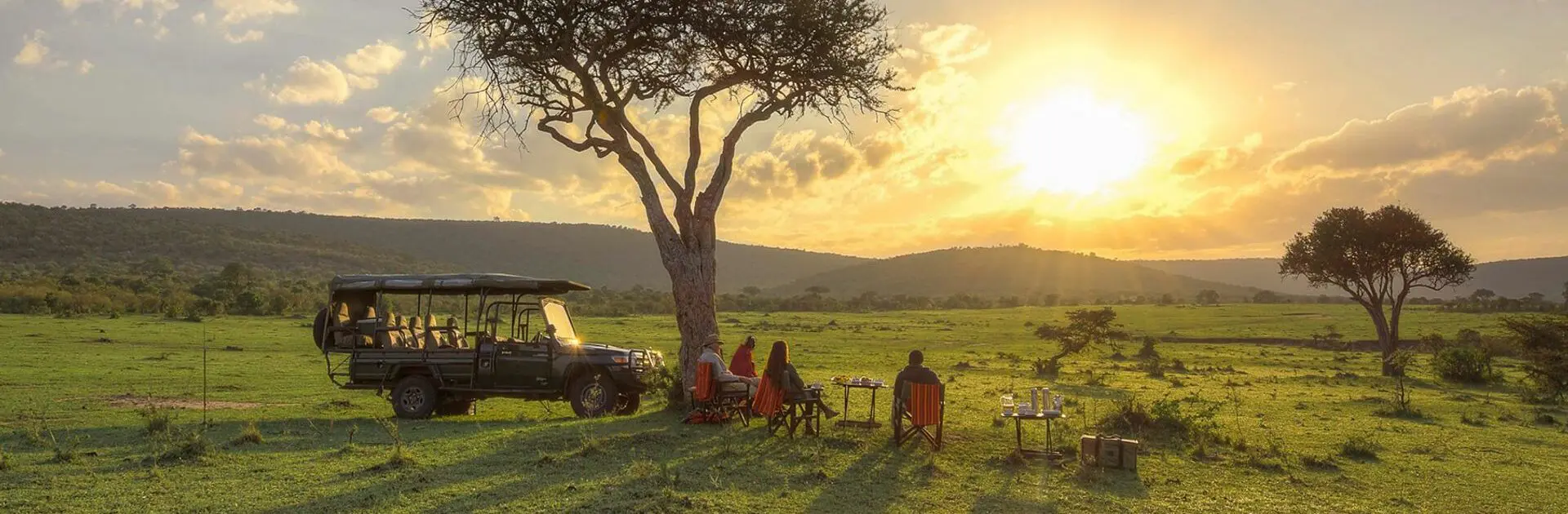 Coucher de soleil sur la savane africaine avec un groupe de personnes assises sous un arbre, un véhicule safari à proximité, et des tables avec des boissons et des objets, dans un paysage ouvert avec quelques autres arbres et des collines à l'horizon.