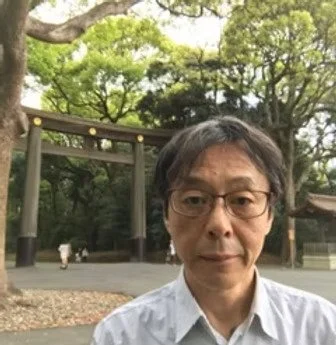 Un homme portant des lunettes et une chemise blanche dans un parc avec des arbres et une structure en bois en arrière-plan.