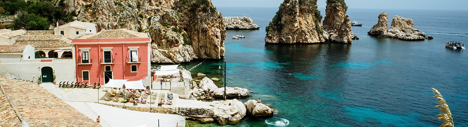Vue panoramique d'une côte méditerranéenne avec des rochers, une plage aménagée avec des parasols, une maison rose, et la mer bleue avec des bateaux.