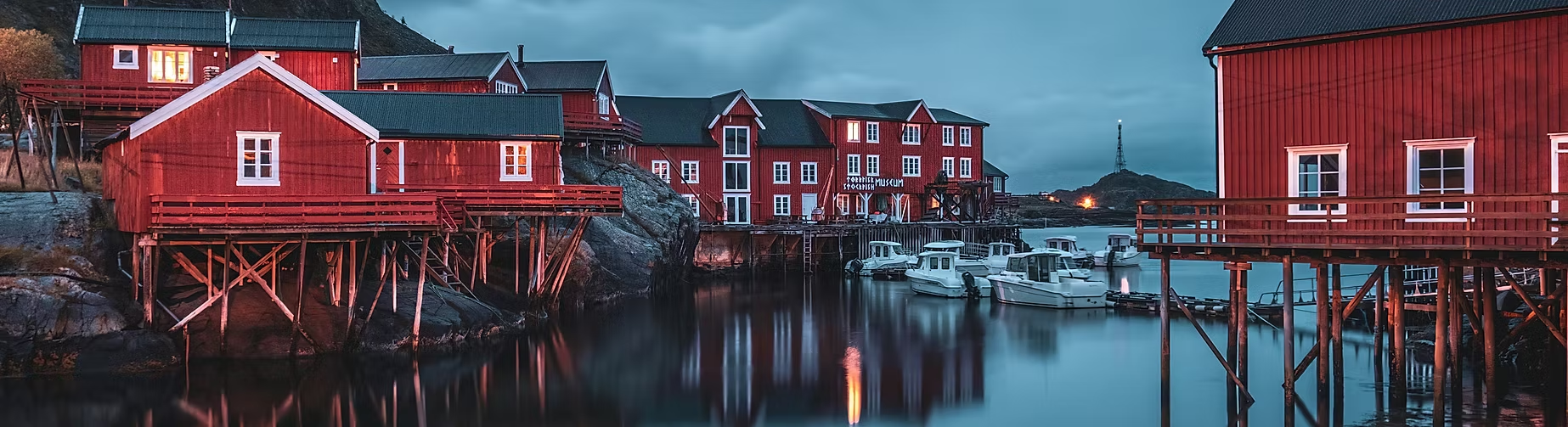 Bâtiments en bois rouges sur pilotis le long d'un quai avec des bateaux amarrés, au bord d'un fjord ou d'une baie, pendant la tombée de la nuit.