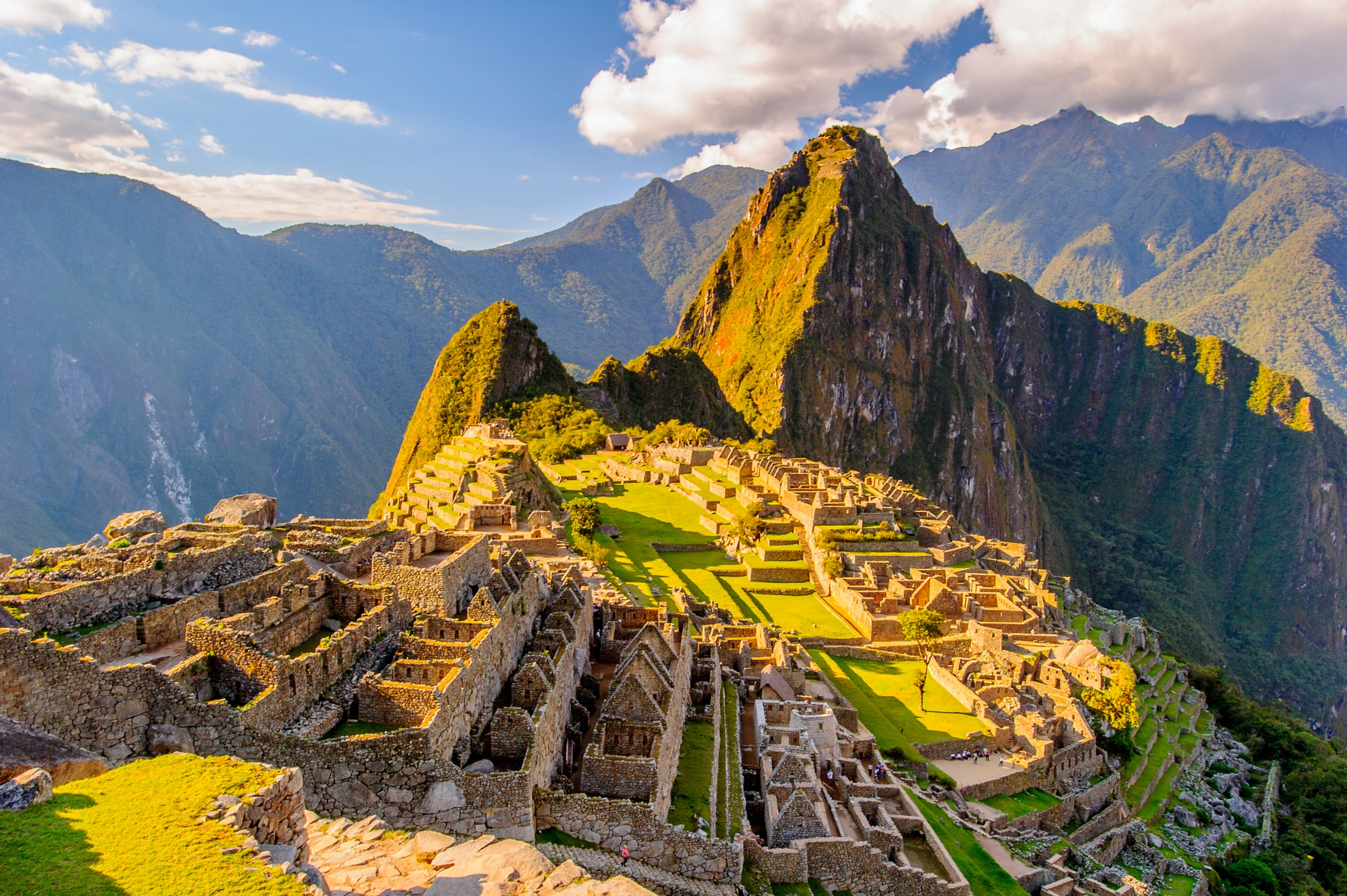 Vue panoramique du site archéologique de Machu Picchu avec des ruines en pierre, des terrasses agricoles, entouré de montagnes verdoyantes sous un ciel partiellement nuageux.