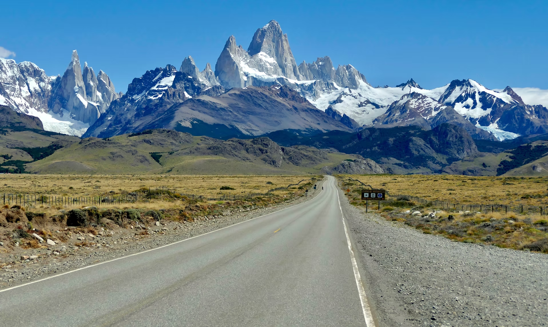 Une route traverse un paysage de plaines avec des montagnes enneigées en fond, sous un ciel bleu clair.
