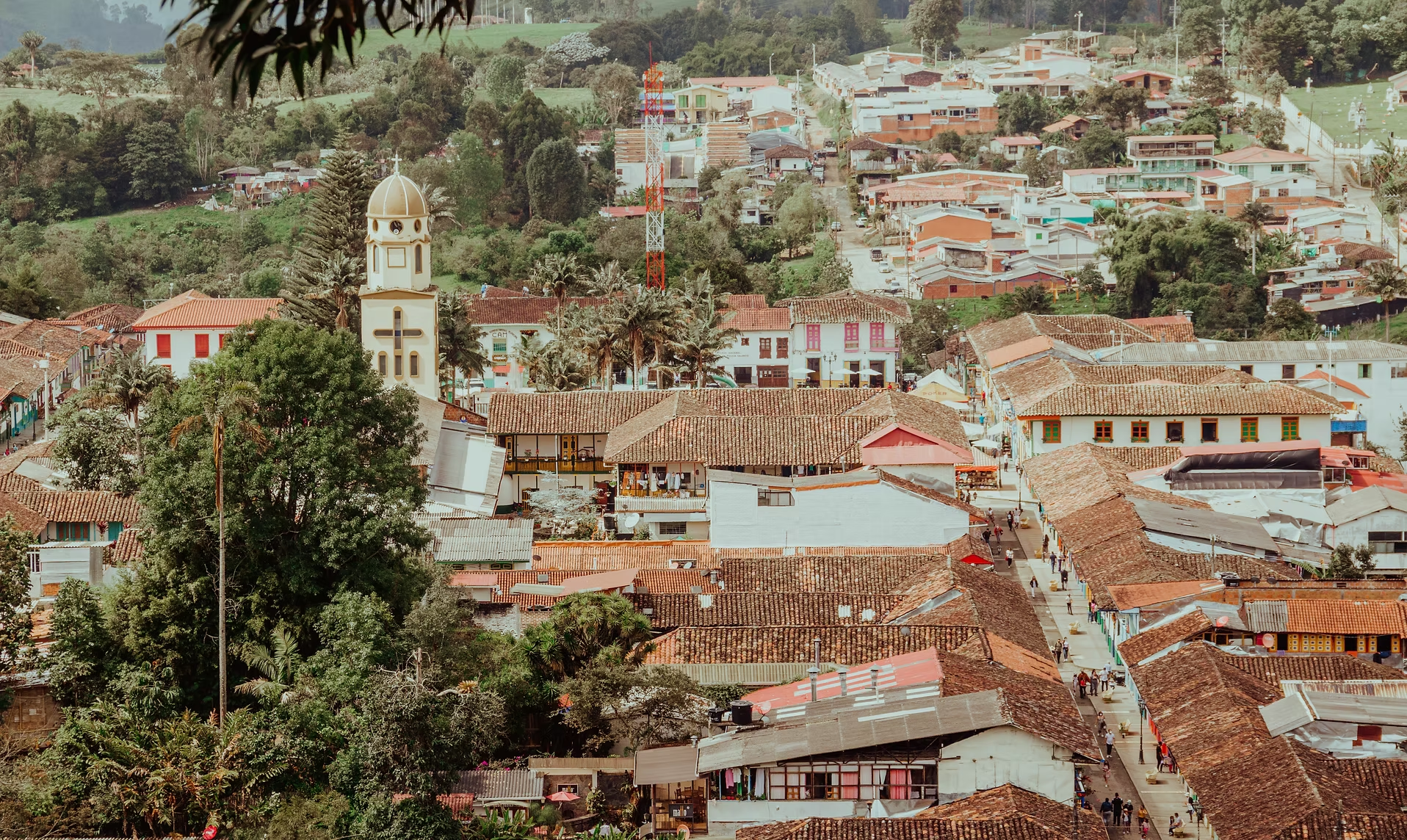 Vue aérienne d'un village avec des toits en tuiles rouges, un clocher d'église, des bâtiments colorés et des rues animées entourées de collines verdoyantes.