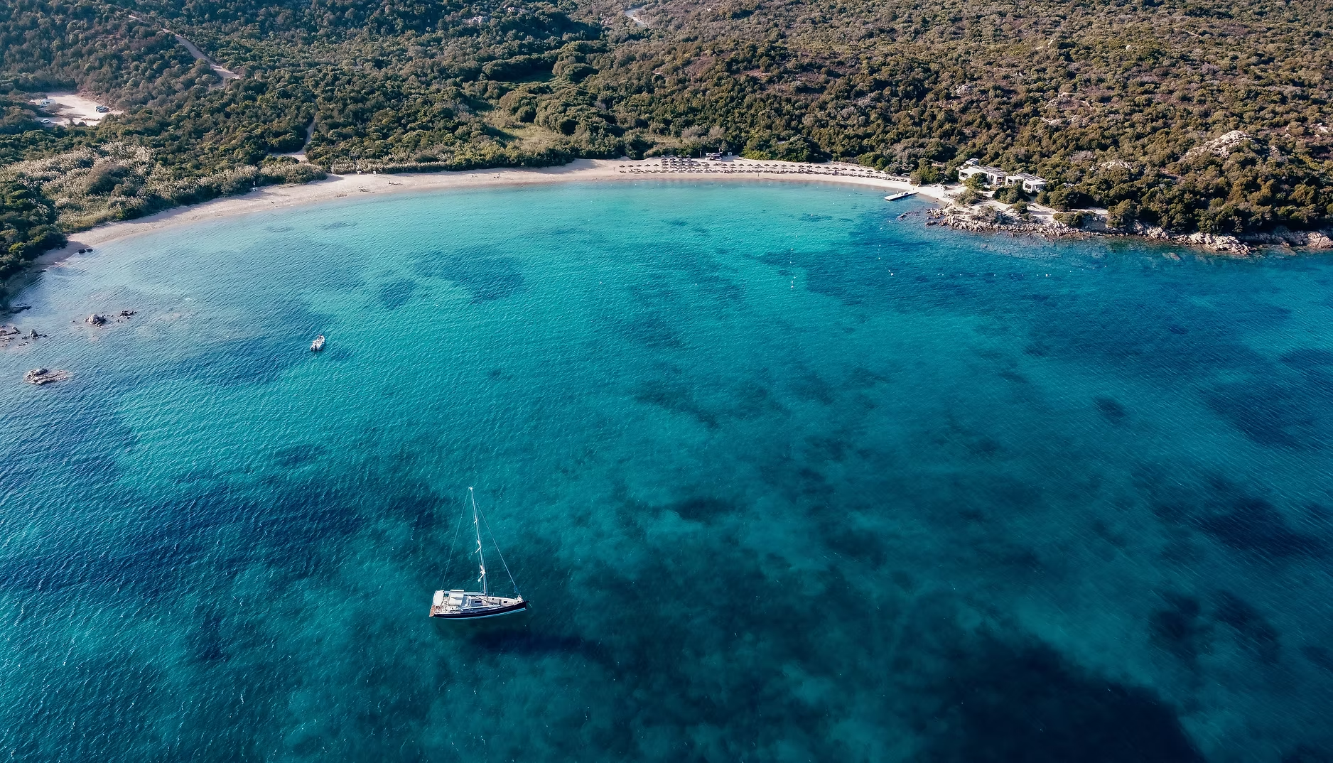 Vue aérienne d'une plage avec une mer turquoise, un bateau à voile dérivant dans l'eau, et une forêt dense en arrière-plan.