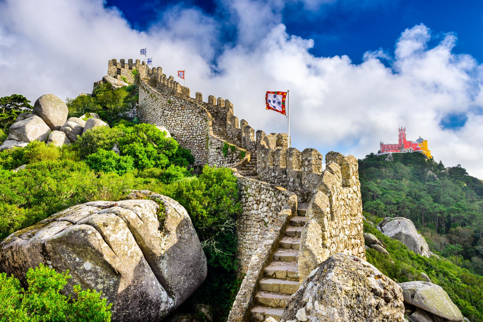 Un sentier en pierre mène à un château à l'arrière-plan, avec des drapeaux et un ciel bleu avec quelques nuages.