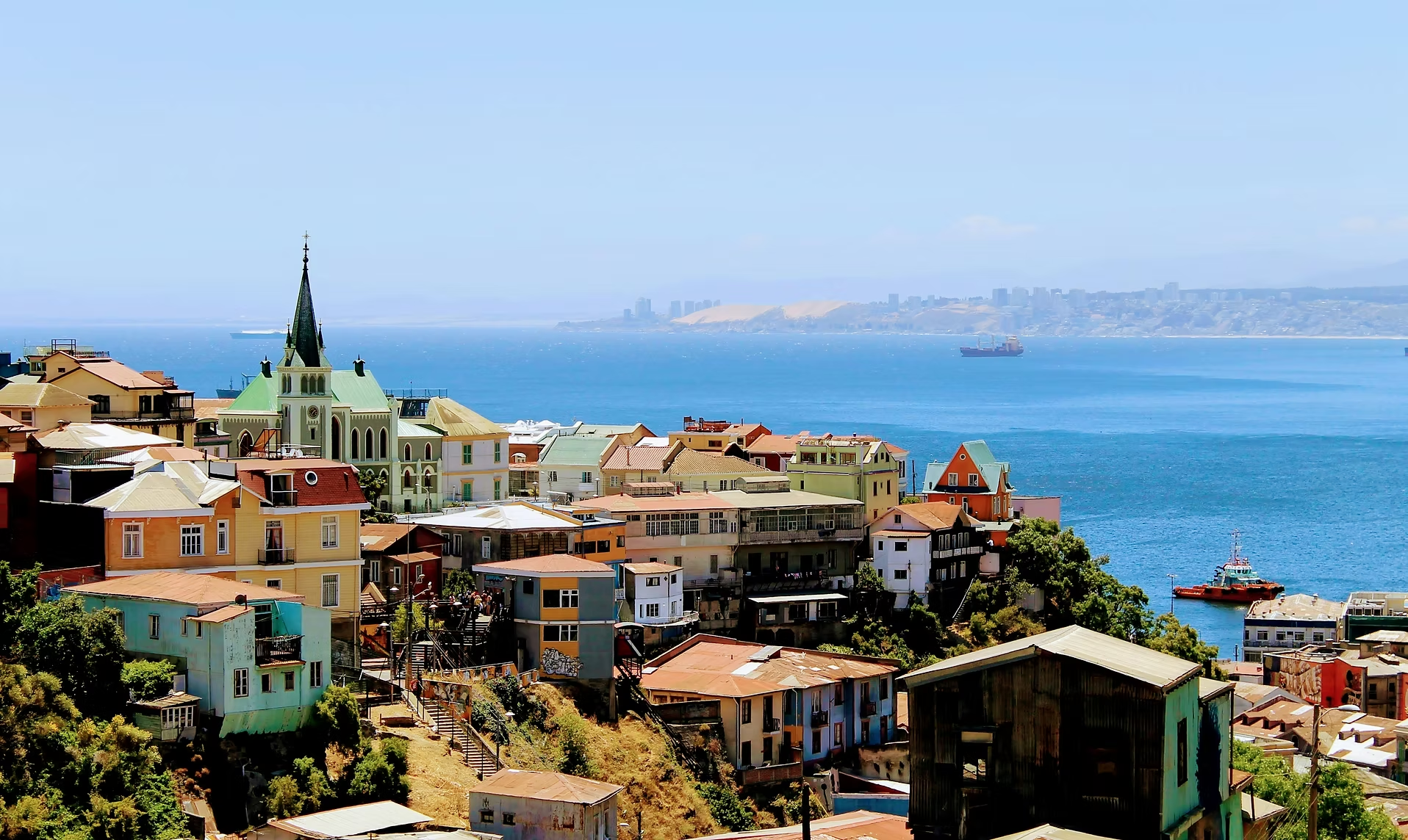 Vue d'une ville côtière avec des bâtiments colorés, un parlement et un port avec des navires, sur fond de mer et de collines au loin.