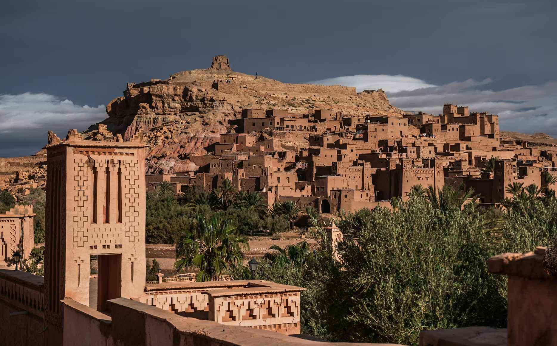 Une vue d'une ville en terre battue avec des constructions en adobe, entourée de palmiers, avec une colline en arrière-plan surmontée d'une ancienne forteresse ou kasbah.