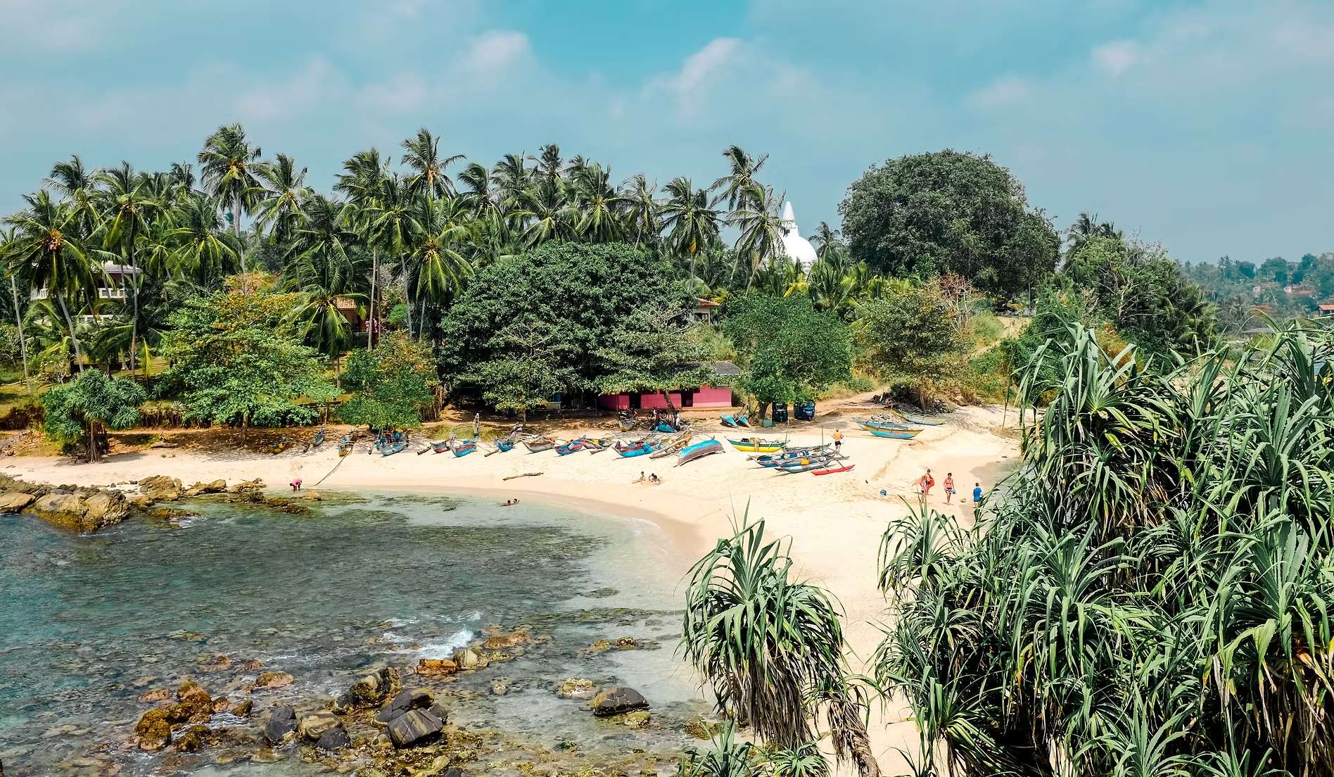 Une plage tropicale avec des bateaux colorés au bord de la mer, entourée de palmiers et de végétation luxuriante, sous un ciel bleu avec quelques nuages.