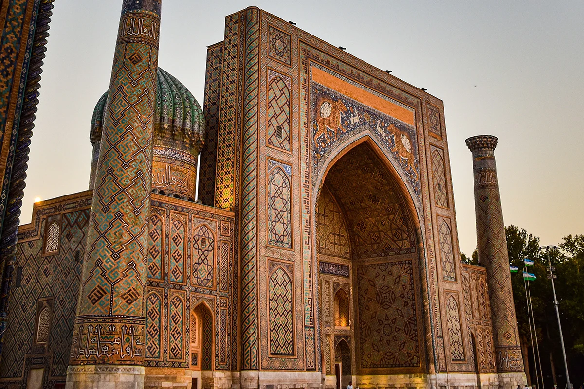 Un bâtiment historique avec une architecture islamique, décoré de mosaïques colorées, comprenant une grande entrée en arche, un minaret et un dôme, probablement situé dans une région d'Asie Centrale ou Moyen-Orient.