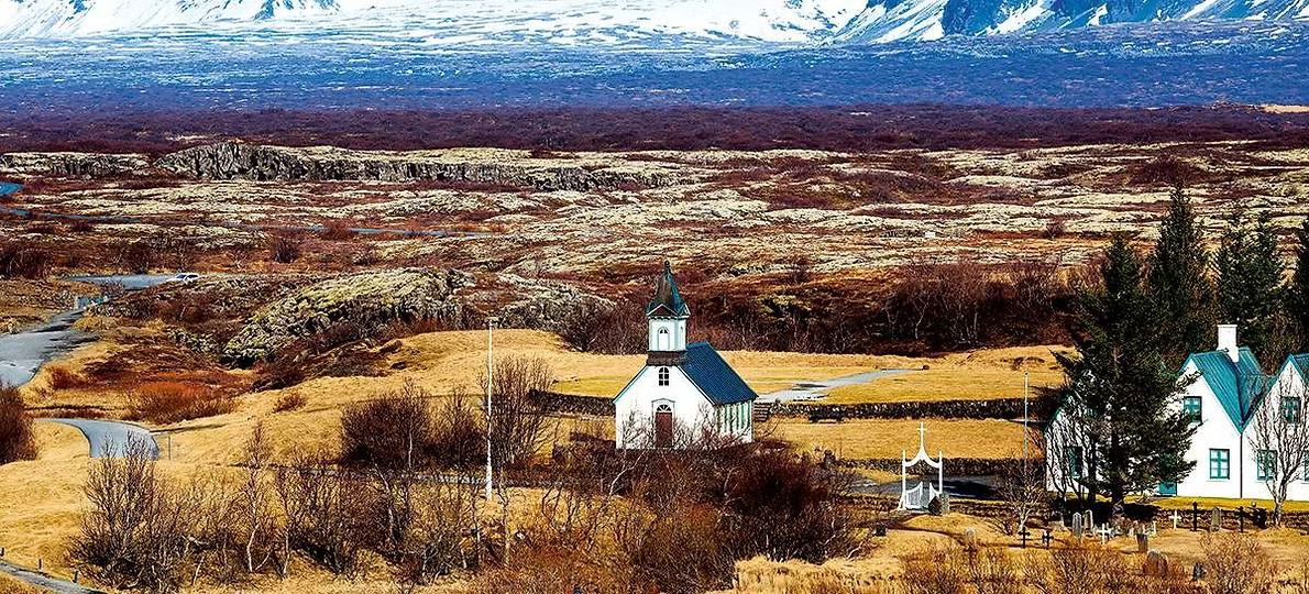 Paysage rural avec une petite église blanche, un bâtiment résidentiel, et des arbres, montagnes enneigées en arrière-plan.