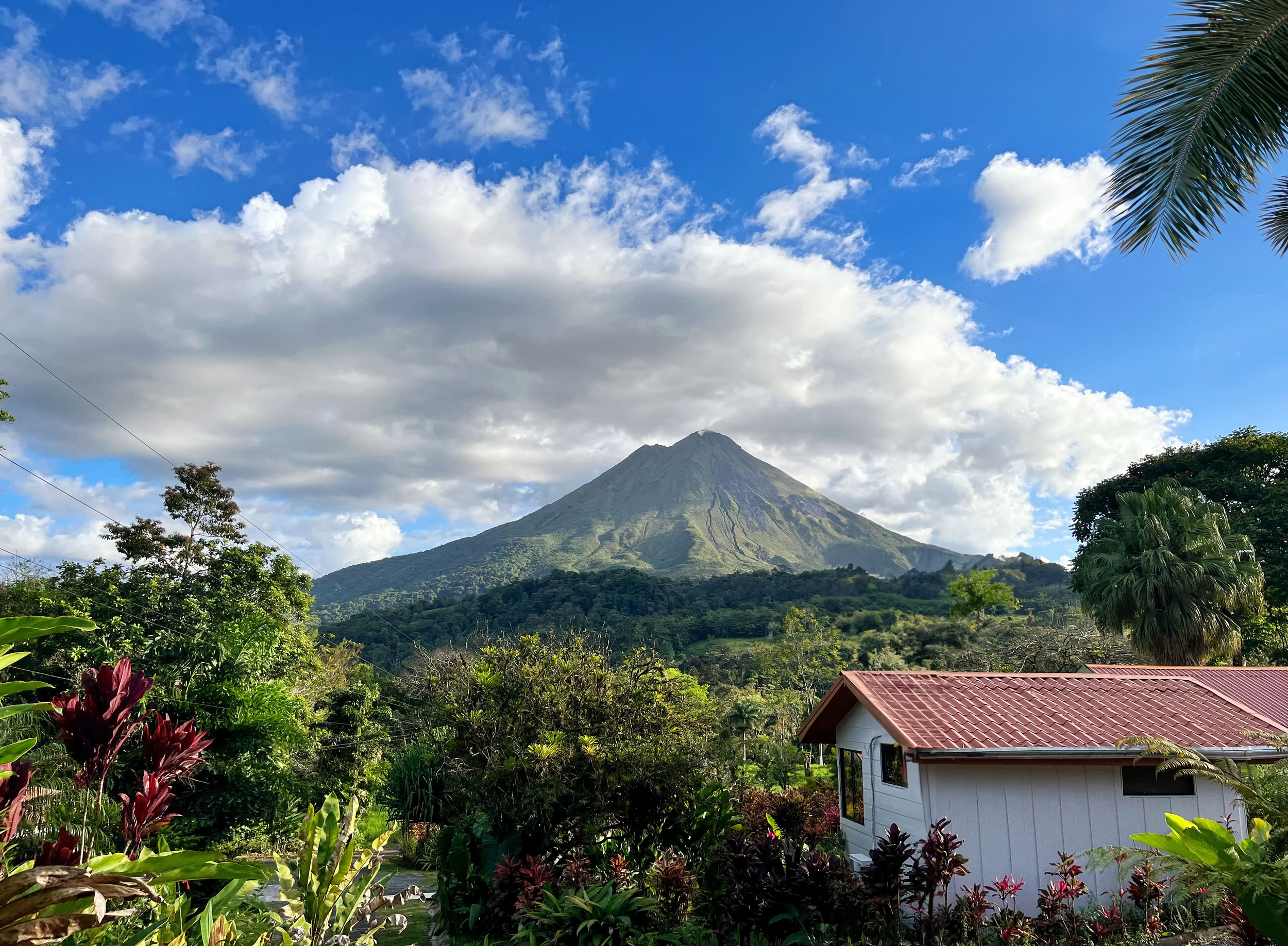 Un volcan en arrière-plan, entouré de nuages et de ciel bleu, avec une végétation dense et une maison à toit rouge au premier plan.