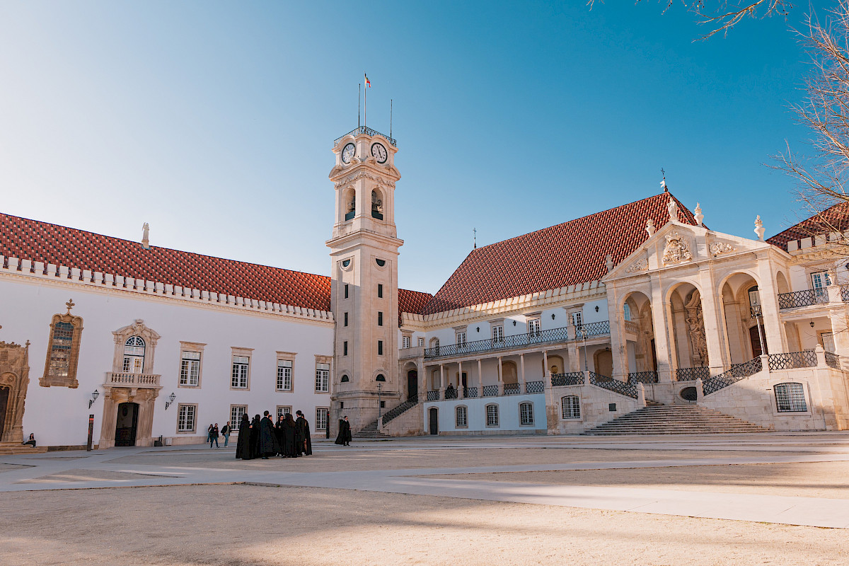 Une photo d'un bâtiment historique avec un clocher, des escaliers et une façade ornée, sous un ciel bleu, avec des personnes en habits traditionnels