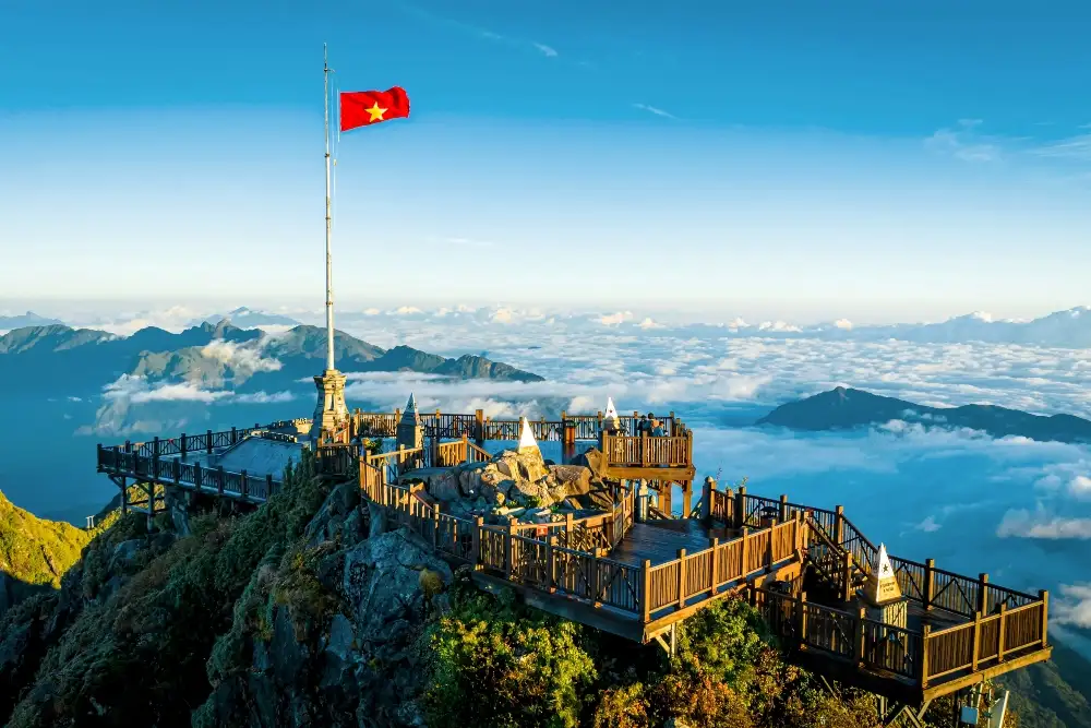 Un drapeau vietnamien flottant sur un mât en haut d'une montagne, avec une plateforme en bois et des montagnes brumeuses en arrière-plan.