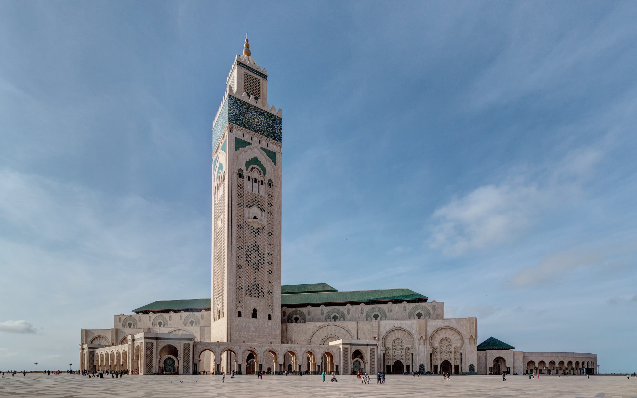 Une grande mosquée avec une minaret ornée de mosaïques colorées, façade en pierre claire, avec un ciel bleu en arrière-plan et des personnes se promenant dans la cour.