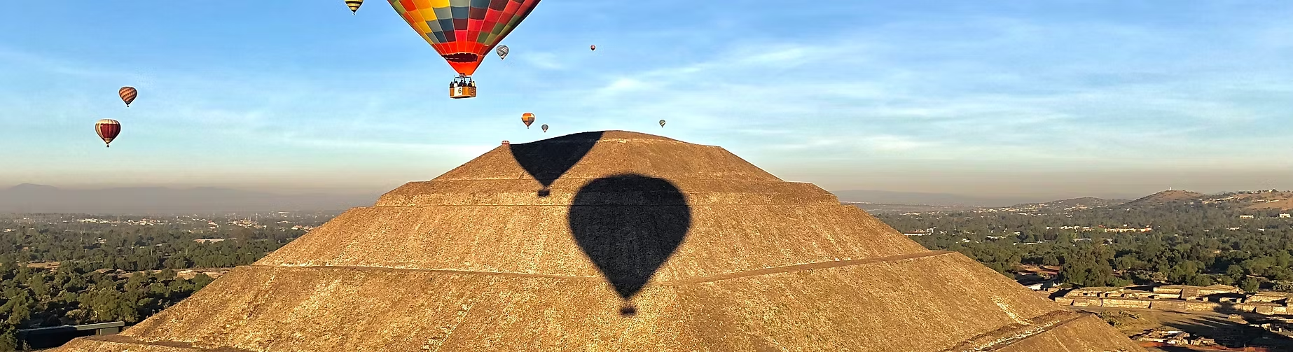 Ballons de plusieurs couleurs volant dans le ciel au-dessus d'une pyramide rocheuse. La pyramide projette une ombre en forme de ballon sur sa surface.
