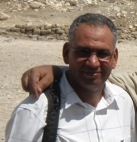 Homme portant des lunettes de soleil, une chemise blanche, dans un environnement désertique, avec une autre personne dont l'épaule repose sur lui.