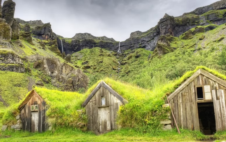 Trois petits bateaux en bois avec des toits en herbe, situés à la base d'une montagne verte avec cascades et un ciel nuageux.