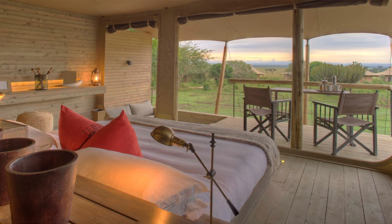 Chambre d'hôtel ou de lodge avec vue panoramique sur la savane ou la campagne, lit avec oreillers rouges, terrasse extérieure avec chaises et table, décor en bois naturel.