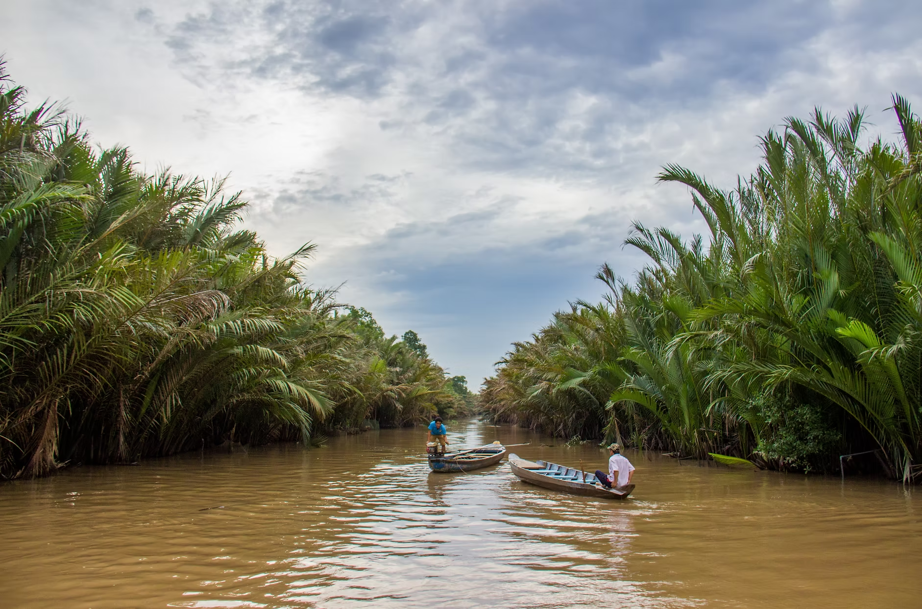 Deux personnes dans des petites barques sur une rivière bordée de palmiers, avec un ciel nuageux au-dessus.