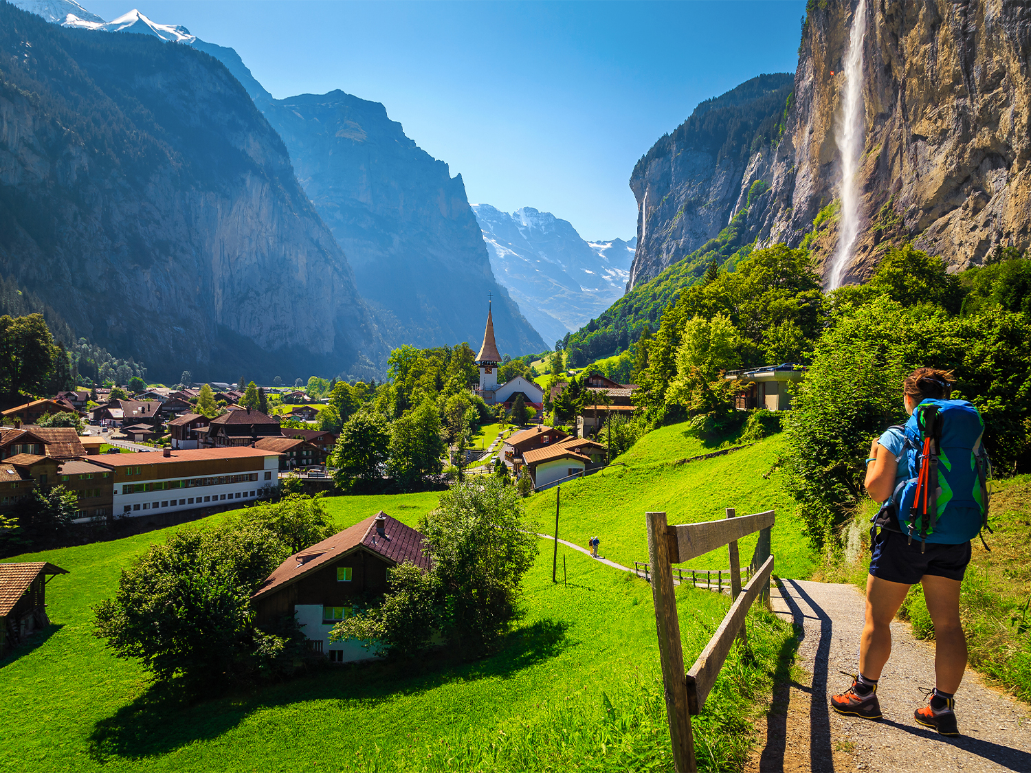 Paysage de montagnes avec une vallée verdoyante, une petite église, des maisons en bois, et une cascade visible sur la paroi rocheuse, avec une personne en randonnée.